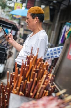 Xian, China -  July 2019 : Bored food seller checking his smartphone on a stall in Muslim Quarter in Xian town, Shaanxi Province