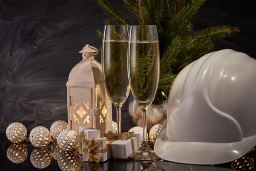 Construction hard hat, wineglasses with champagne, gift boxes and Christmas ornament on black background. Construction New Year, Christmas background with copy space. For greeting card or advertising
