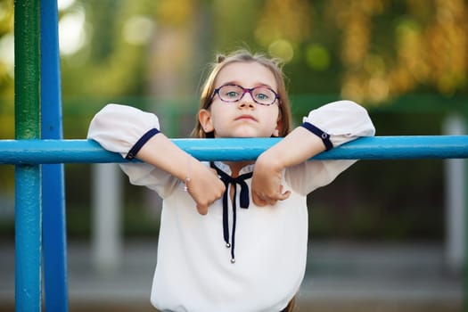 Pensive child girl leaning on the horizontal bar. Schoolgirl with glasses looking at the camera. Selective focus.