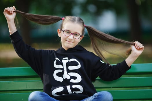 Girl with hairstyle ponytails sitting on a bench. Child with long hair. Selective focus.