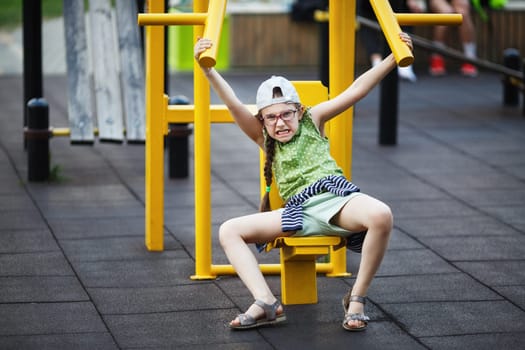 Angry little girl making exercises on sports training apparatus outdoors. Child goes in for sports and shakes muscles. Selective focus.