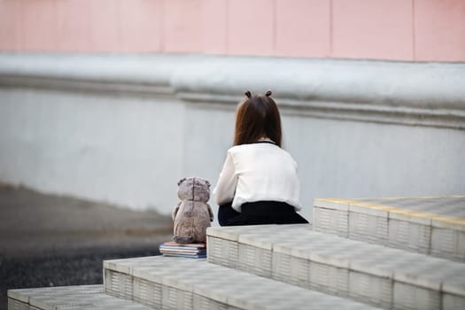 Sad girl with a plush toy sits on the steps of the school stairs with her back to the camera. Offended child. Selective focus.