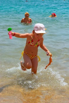 Happy little girl running out of sea. Positive childish emitions. Child running away from sea water and holding doll in hand. Happy childhood. Baby girl having fun with toys in sea during family vacations