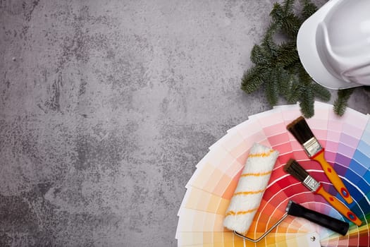 Construction hard hat, color palette guide, paint brushes and roller, fir tree branches on grey background. Top view with copy space, New Year and Christmas. Concept of construction or design office