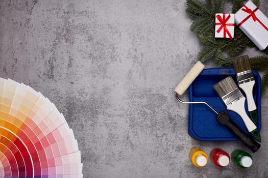 Color palette guide, paint brushes, roller, cans of paint, fir tree branch and gift boxes on grey background. Top view, copy space, New Year and Christmas. Concept of construction or design office