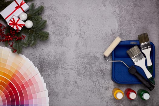 Color palette guide, paint brushes, roller, cans of paint, fir tree branch and gift boxes on grey background. Top view, copy space, New Year and Christmas. Concept of construction or design office