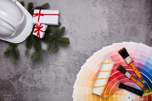 Construction hard hat, color palette guide, paint brushes and roller, fir tree branches and gift boxes on grey background. Top view with copy space, New Year and Christmas. Concept of construction or design office