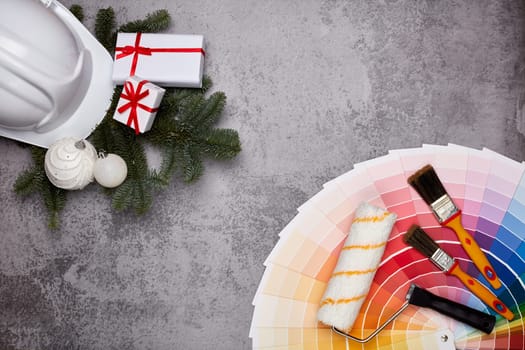Construction hard hat, color palette guide, paint brushes and roller, fir tree branches and gift boxes on grey background. Top view with copy space, New Year and Christmas. Concept of construction or design office