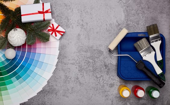 Color palette guide, paint brushes, roller, cans of paint, fir tree branch and gift boxes on grey background. Top view, copy space, New Year and Christmas. Concept of construction or design office