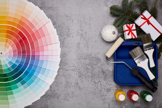 Color palette guide, paint brushes, roller, cans of paint, fir tree branch and gift boxes on grey background. Top view, copy space, New Year and Christmas. Concept of construction or design office