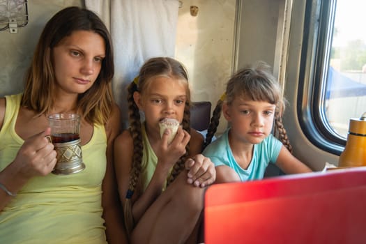 Mom and two girls are looking at the laptop screen in the train car, having a bite while