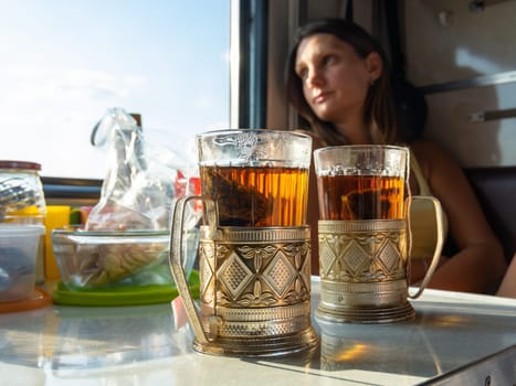 On the table in the compartment of the train are glasses with tea in metal cup holders, in the background the girl looks out the window