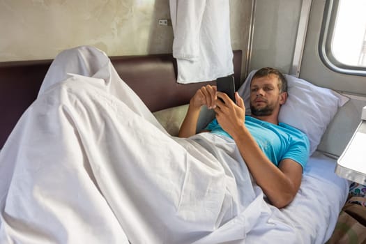 A man lies on the bottom shelf in a train car with a phone in his hands