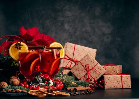Still life with glasses of mulled wine or fruit tea, Red Poinsettia flowers, fir branch and gift boxes on a dark background with copy space. New year and Christmas table setting. Greeting card.