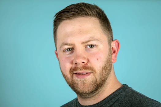 Bearded casual young man in a grey t-shirt looking pensively at the camera in a close up frontal studio portrait