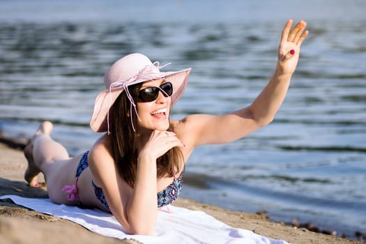 The smiling cheerful woman is greeting, saying hello and waving hand to someone on the beach at summer.