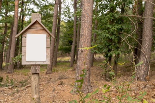 Unlabeled white sign on a wooden board with a small roof. Screwed to a wooden stake in the woods