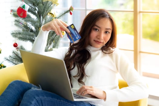 Young Adult teenager using laptop on sofa in living room for online shopping with gift box with Christmas tree in background