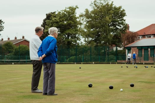 A rear view shot of senior adults spectating a lawn bowling game, standing with their hands behind their back.