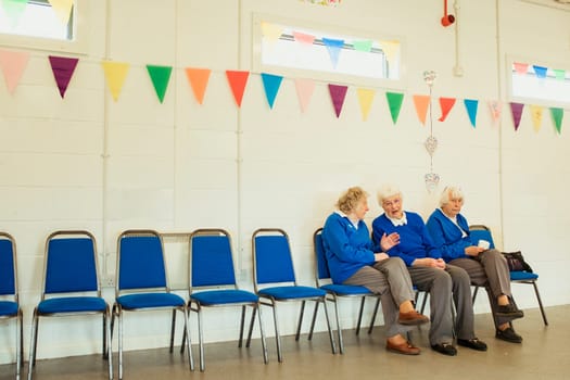 Three senior women talking and sitting indoors on blue chairs in a hall decorated with bunting.