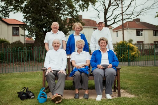 A group of senior adults sitting on a bench at a bowling green, looking at the camera.