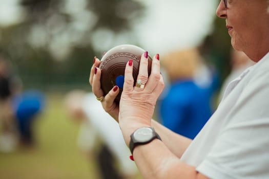 A close up shot of a senior woman holding a bocce ball, ready to take her shot. She is wearing nail polish and a watch.