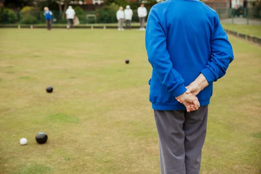 A rear view shot of an unrecognizable person spectating a lawn bowling game, standing with their hands behind their back.