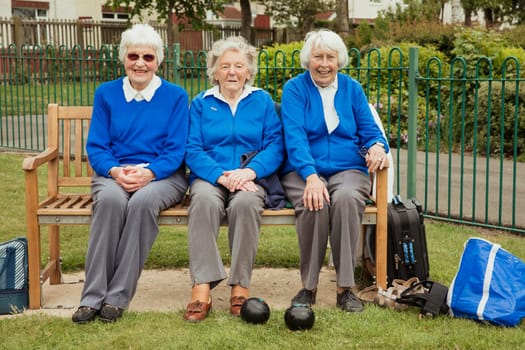 Three senior friends sitting on a bench at a bowling green, relaxing and smiling at the camera.