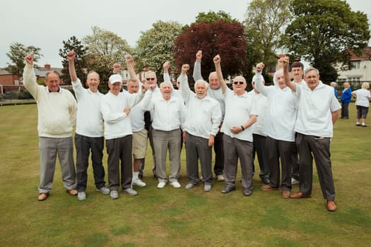 A front view shot of a community of senior men punching the air, celebrating after their lawn bowling game.