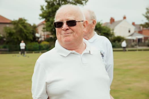 A portrait of a senior man smiling at a bowling green. He is looking away and wearing sunglasses.