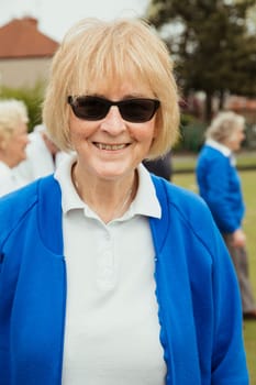 A portrait of a senior woman smiling at a bowling green. She is wearing sunglasses.
