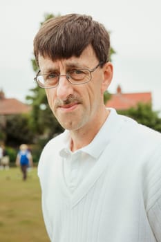 A portrait of a senior man smiling at a bowling green. He is wearing glasses.