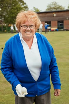 A portrait of a senior woman smiling at a bowling green, holding a bocce ball.