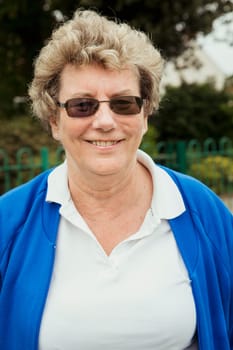 A portrait of a senior woman smiling at a bowling green. She is wearing sunglasses.