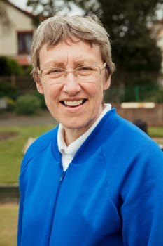 A portrait of a senior woman smiling at a bowling green. He is wearing glasses.