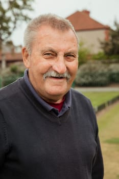 A portrait of a senior man with a moustache smiling at a bowling green.