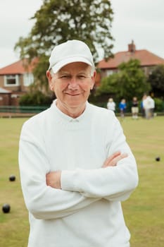 A portrait of a senior man smiling at a bowling green. He is wearing a white cap and is crossing his arms.