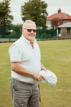 A portrait of a senior man smirking at a bowling green. He is wearing sunglasses and is holding a white cap.