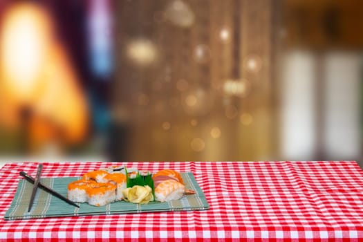 Table top on sushi background. Closeup of various kinds of sushi and chopsticks served on green bamboo mat on a red checkered tablecloth against abstract blurred light brown background. Space.