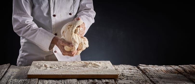 Baker of chef kneading a large portion of dough with his hands during preparation over a floured wooden board in a rustic panorama banner with copy space