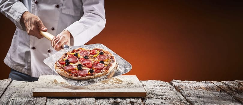 Chef in a pizzeria with homemade salami pizza displayed on a metal paddle over a floured board in front of the glow of the fire in the oven in a rustic panorama banner with copy space