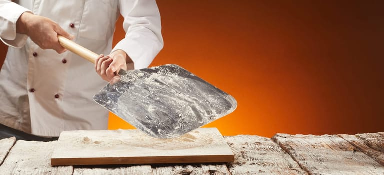 Chef holding a floured empty metal pizza paddle over an old rustic or vintage wooden kitchen table for food placement in a panorama banner with copy space