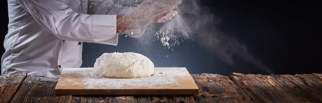 Panorama banner of a chef clapping his hands with a cloud of flour in the air as he dusts a mound of uncooked pastry on a board while making pizzas or baking