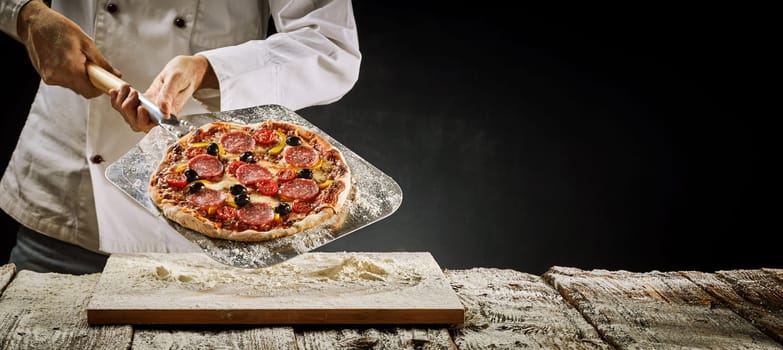 Chef removing an Italian salami pizza from the oven displaying it on a metal paddle over a floured wooden board in a rustic panorama banner with copy space