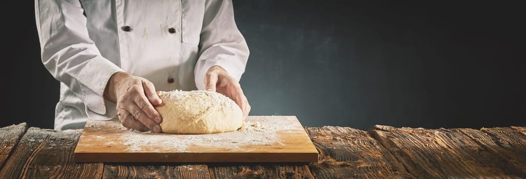 Chef in a pizzeria or baker preparing a mound of raw dough on a floured wooden board on a rustic table in a panorama banner with copy space