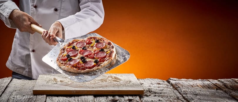 Chef with a delicious homemade pepperoni pizza displaying it on a metal paddle after removing it from the oven in a pizzeria in a rustic panorama banner with copy space with glow of the fire