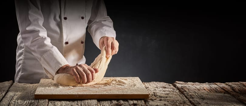 Hands of a male chef, cook or baker kneading dough on a cutting board over on old rustic wooden kitchen table in a rustic panorama banner with copy space