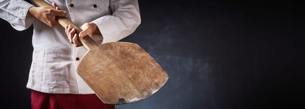 Chef holding an empty wooden pizza paddle in a close up on his hands in a panorama with dark background and copy space for food or product placement