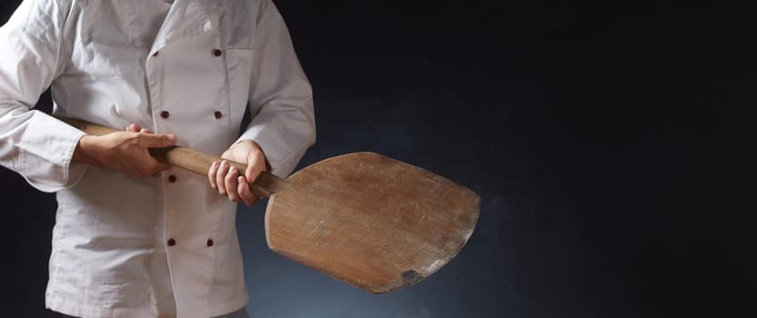 Chef in white uniform holding an empty wooden pizza paddle with placement copy space for food over a dark background