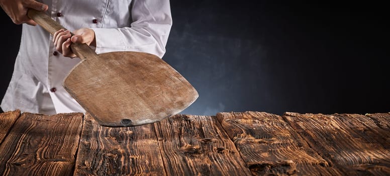 Chef holding an empty wooden pizza paddle over an old rustic kitchen table in a panorama banner with copy space for product or food placement
